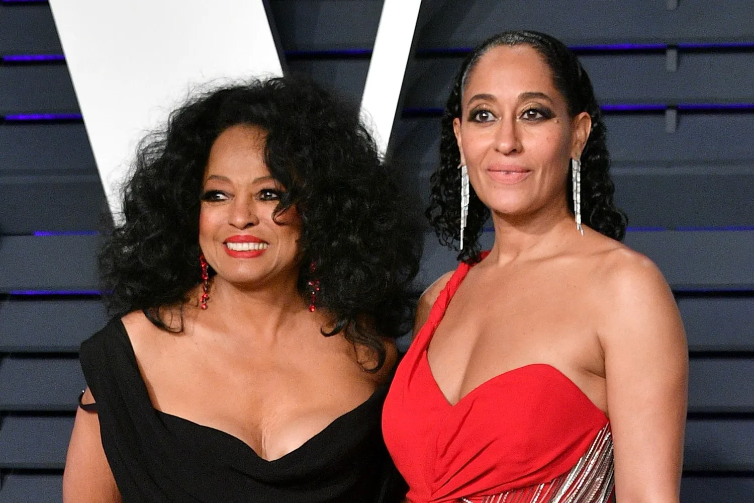 Two women in elegant evening gowns pose together, smiling at an event.