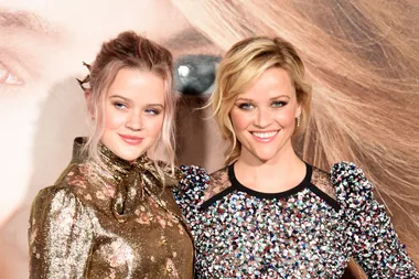 Two women smiling at an event, wearing sparkly outfits with a backdrop featuring a large eye and hair.