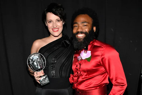 Two people smiling, one holding a BAFTA award, against a black background.