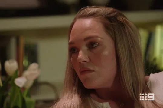 A woman with long hair looks to the side in a dimly lit room with flowers in the background; Nine Network logo visible.