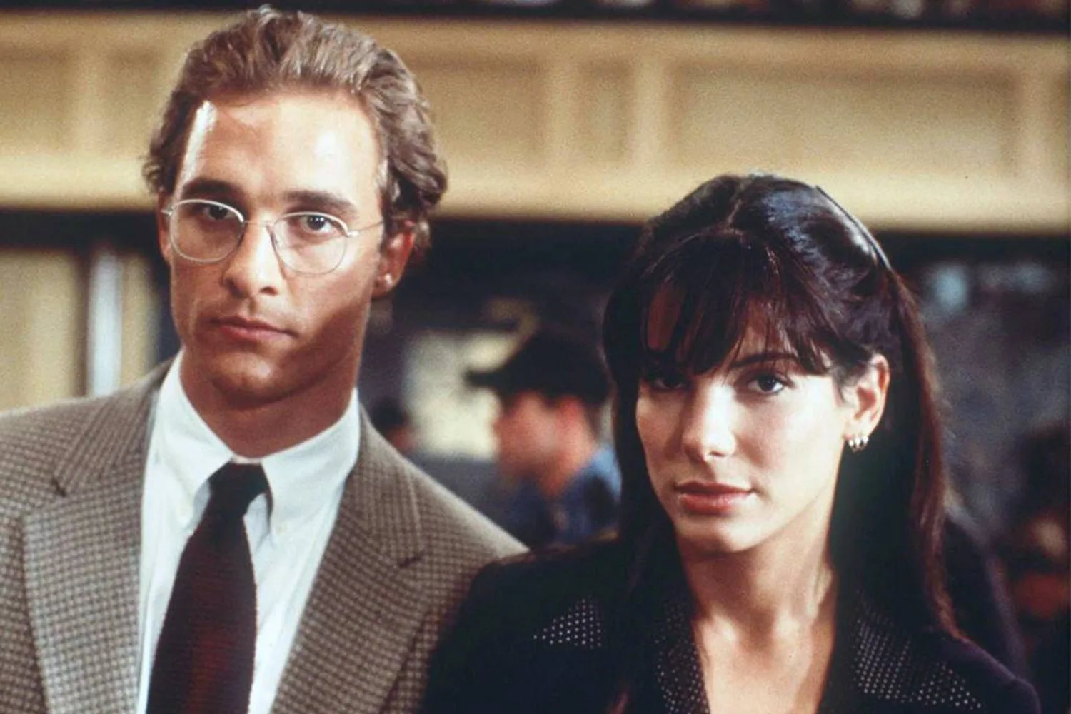 Two people standing side by side, one wearing glasses and a suit, the other with long hair and earrings, in a formal setting.