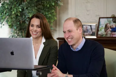 A man and woman smiling in front of a laptop, sitting in a room with greenery and framed photos in the background.