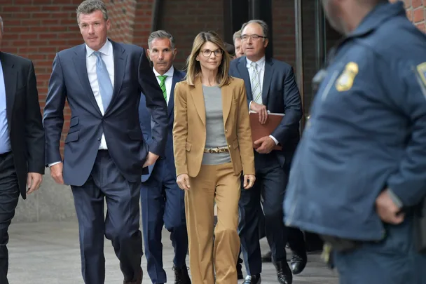 A woman in a tan suit walks with a group of men in suits outside a building, guarded by security.