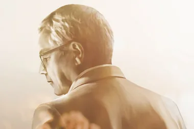A man in glasses and a suit, facing away in a sepia-toned image with soft lighting.