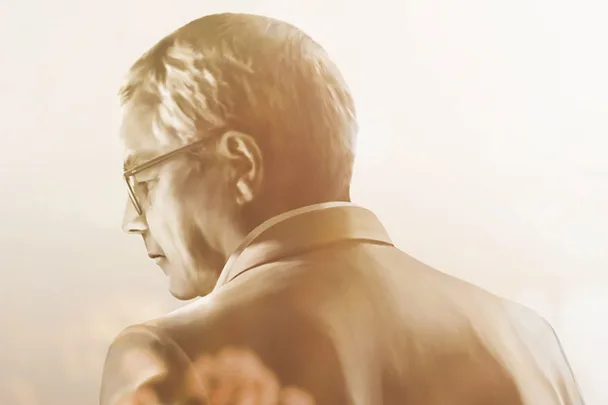 A man in glasses and a suit, facing away in a sepia-toned image with soft lighting.