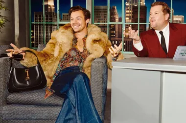Two people sitting and smiling on a talk show set, one wearing a fur coat and floral shirt, the other in a red suit.