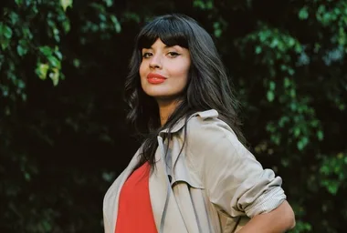 A person with long dark hair in a trench coat and red top stands outdoors with greenery in the background.