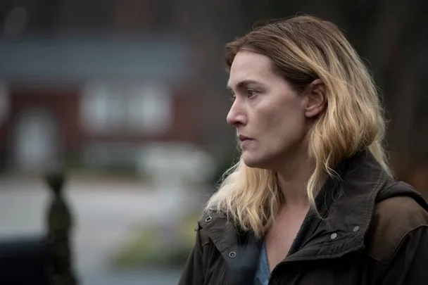 A woman with wavy blonde hair in a somber setting, wearing a dark coat, looks thoughtfully into the distance.