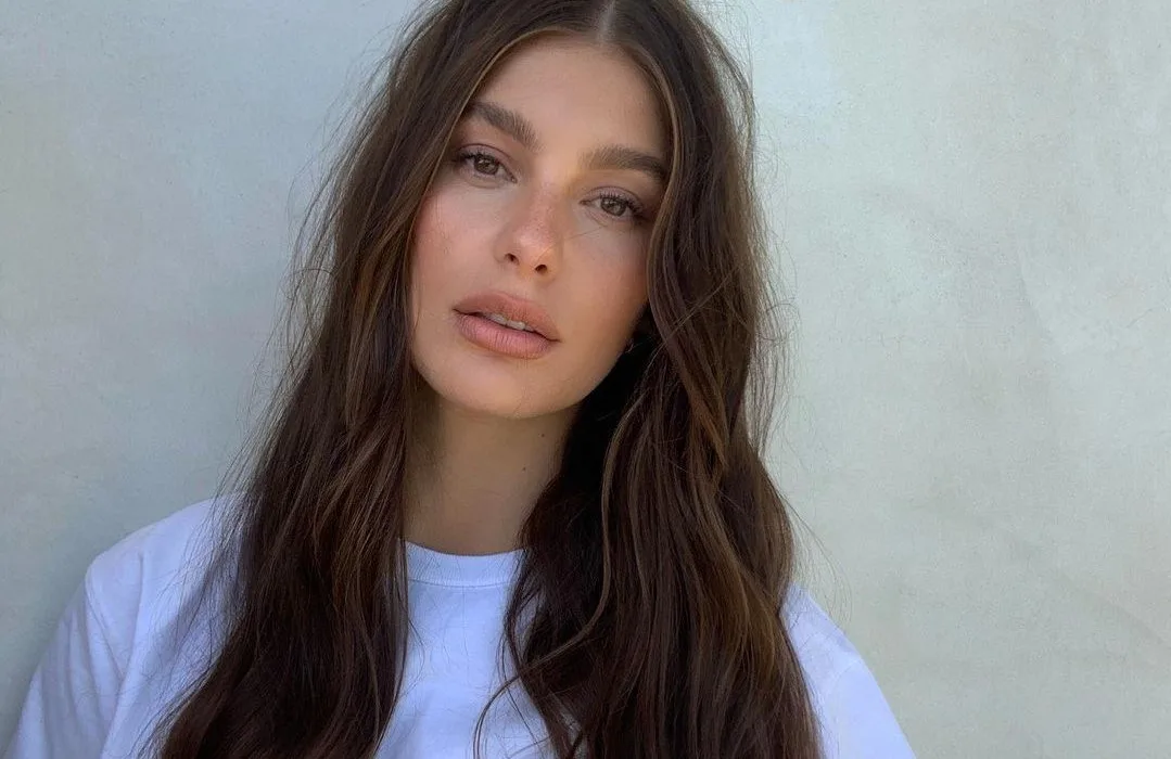Young woman with long brown hair and a white shirt against a light gray background.