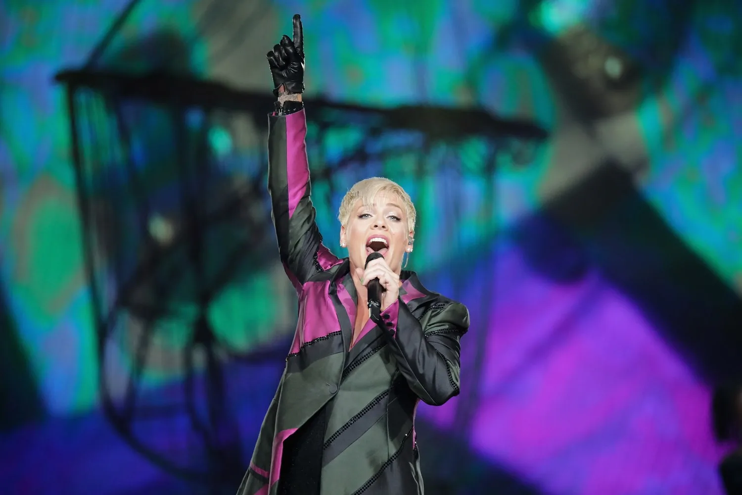 Singer in a pink and black outfit performs energetically on stage, holding a microphone and raising one arm.