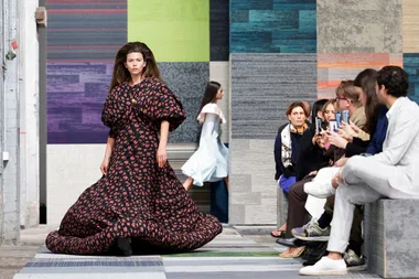 Model in a puffy black and red gown walks runway, audience capturing photos with phones.
