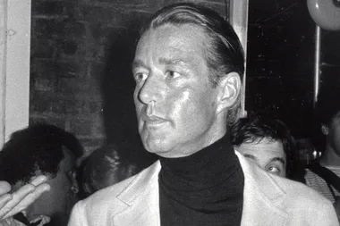 A man in formal attire stands in a dimly lit room, looking to his right, with others in the background.