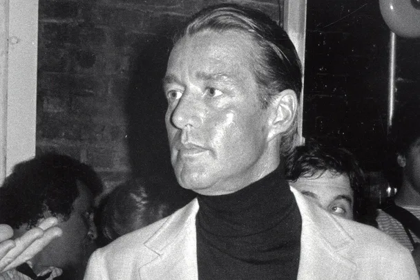 A man in formal attire stands in a dimly lit room, looking to his right, with others in the background.