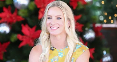 Blonde woman in a yellow floral dress smiling, with a holiday-themed background of red flowers and ornaments.