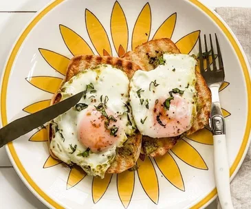 Two fried eggs on toast with herbs on a yellow sunflower plate, knife on left, fork on right.