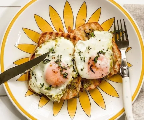 Two fried eggs on toast with herbs on a yellow sunflower plate, knife on left, fork on right.