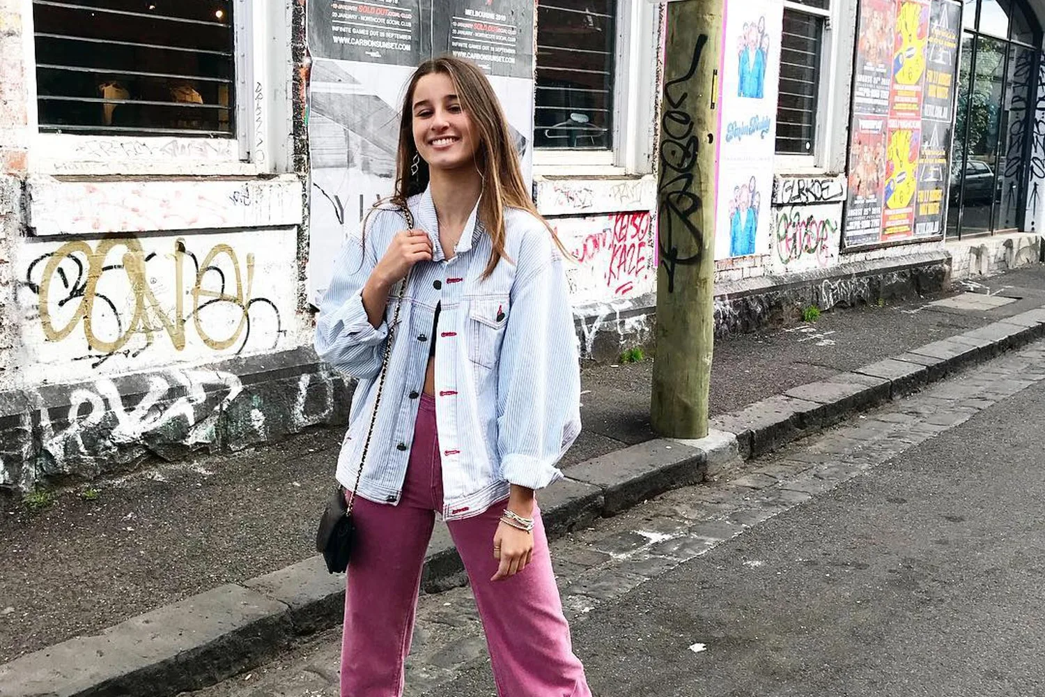 A woman wearing a denim jacket and pink pants smiles on a graffiti-decorated street.