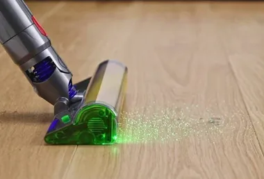 Vacuum cleaner head with green light on hardwood floor, highlighting dust particles.