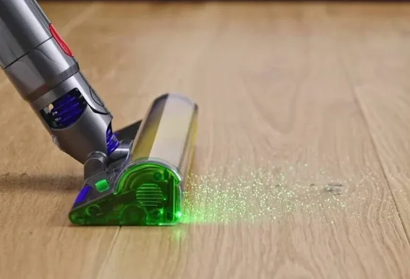 Vacuum cleaner head with green light on hardwood floor, highlighting dust particles.