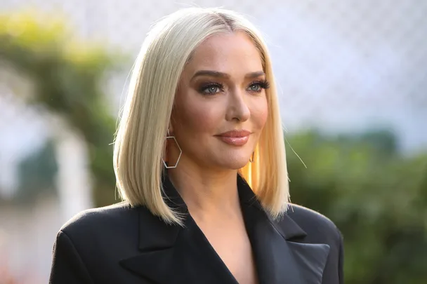 Blonde woman with bob haircut and geometric earrings, wearing a dark blazer, outdoors with blurred background.