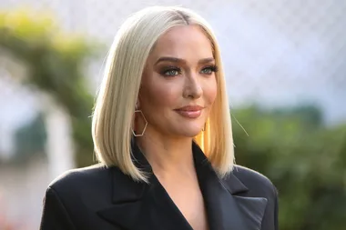 Blonde woman with bob haircut and geometric earrings, wearing a dark blazer, outdoors with blurred background.