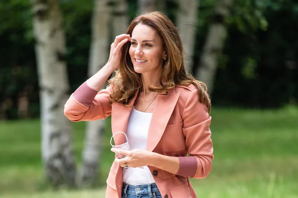 A woman in a pink blazer smiles outdoors, holding a floral face mask in her hand.