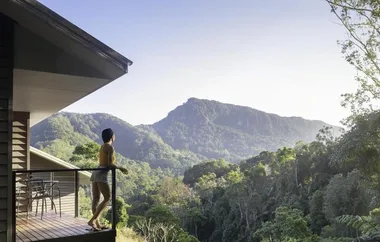 Person standing on a balcony gazing at lush green mountains under a clear sky.