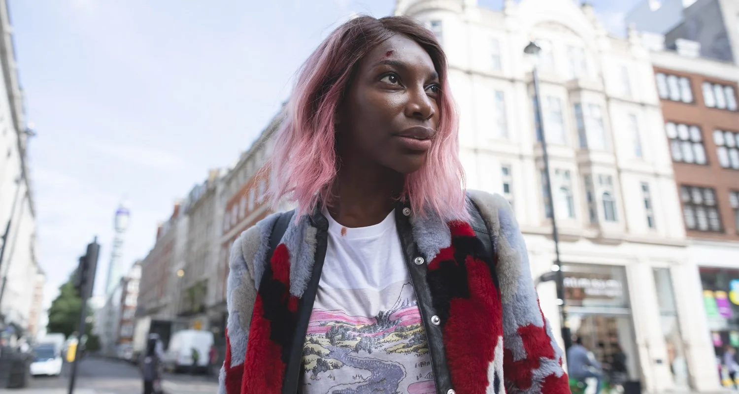 Person with pink hair and patterned coat on a city street, buildings in the background.
