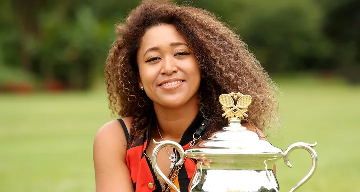 Smiling woman with curly hair holds a large silver trophy outdoors.