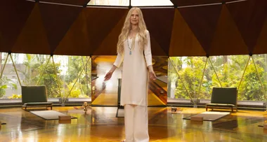 A woman in a white dress stands in a geometric room with large windows and greenery outside.