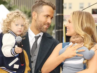 Man in suit with child holding microphone, woman smiling holding a baby, outdoors event setting.