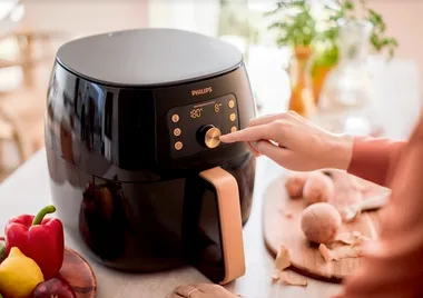 Person adjusting settings on a Philips air fryer, with vegetables and peeled potatoes on the counter.