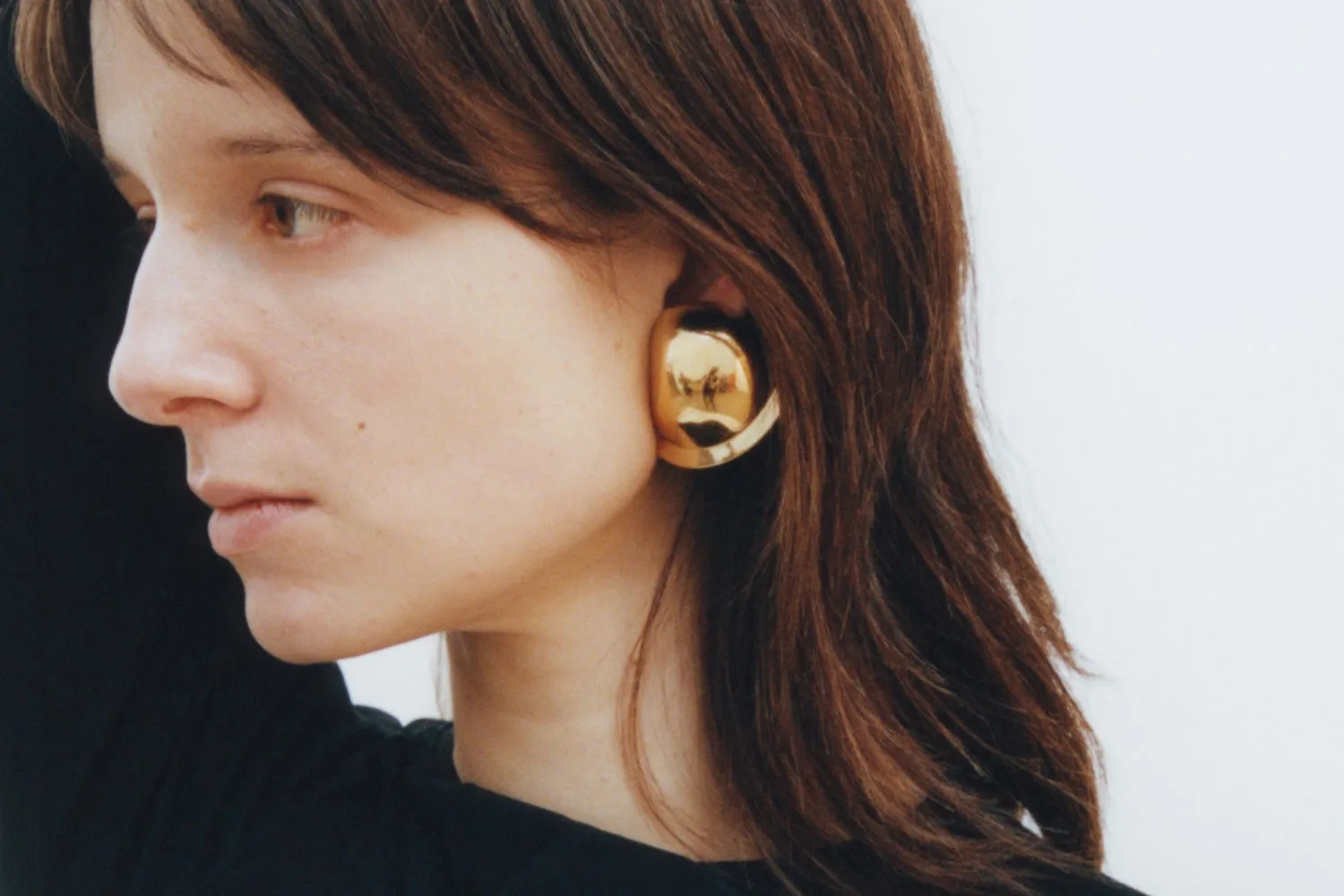 A person wearing large, reflective gold earrings and a black top, looking to the side, with brown hair.