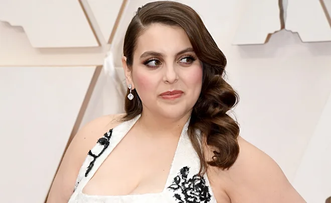 A person with long, dark wavy hair, wearing a white dress with black floral patterns, looks to the side at an event.