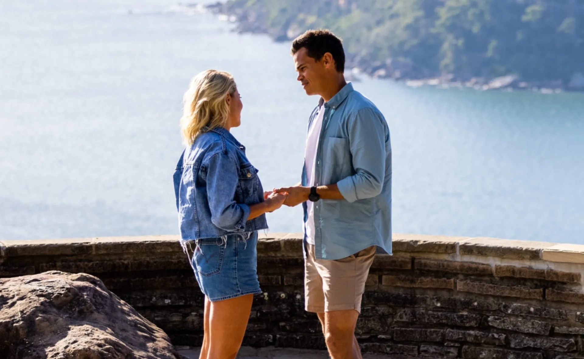 Man and woman holding hands by a scenic ocean view, both wearing casual clothes.