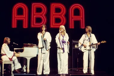 ABBA performing on stage, wearing white outfits with "ABBA" in large red letters behind them.