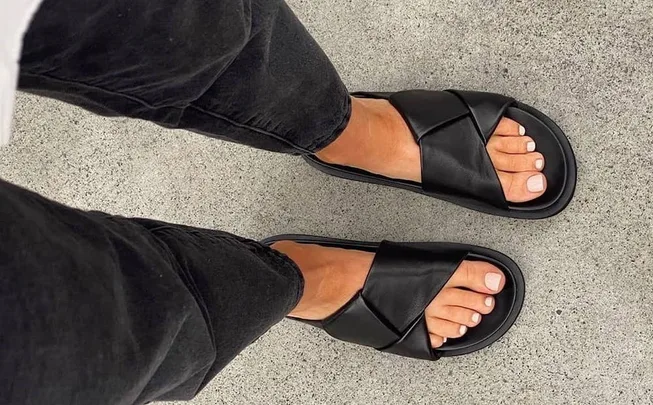 Person wearing black crisscross sandals and black pants on gray concrete floor.