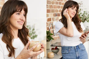 Woman holding a coffee cup and wearing earbuds, standing in a kitchen with a bowl of fruit.