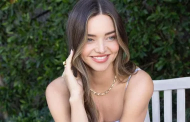 A person smiling seated outdoors, wearing a gold necklace and ring, with green foliage in the background.