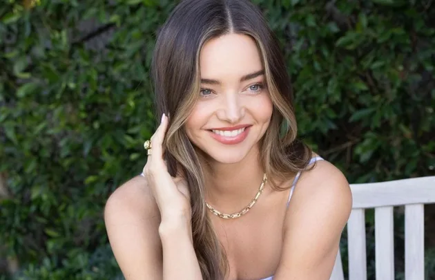 A person smiling seated outdoors, wearing a gold necklace and ring, with green foliage in the background.