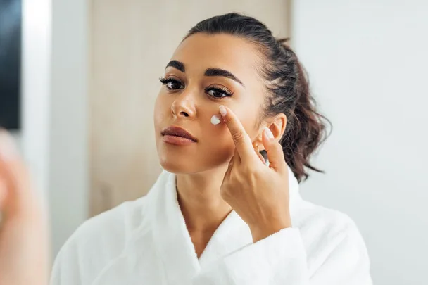 Woman applying facial cream, wearing a white robe, looking in mirror.
