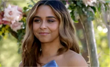 A woman with long hair smiles outdoors, with flowers and greenery in the background.
