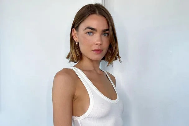 A woman with short brown hair in a white tank top stands against a plain background.