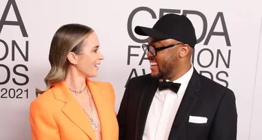 Two people smiling at each other at the CFDA Fashion Awards 2021, one in an orange coat, the other in a tuxedo and cap.