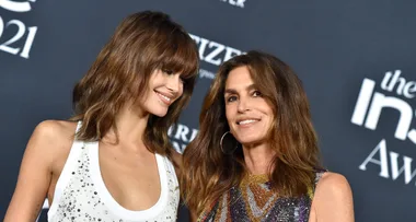 Two women smiling at the 2021 awards event; one in a white dress, the other in a colorful dress, on a blue carpet backdrop.