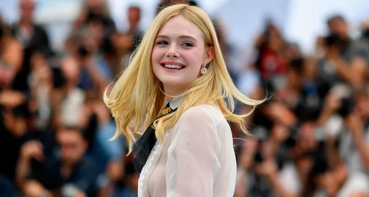 A smiling person with long blonde hair, wearing a sheer blouse, stands in front of a blurred crowd of people.