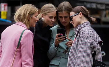 Four fashionable women closely viewing a smartphone together, standing outdoors, enjoying a moment of shared interest.