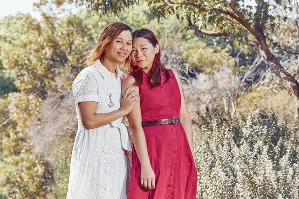 Two people standing outdoors, one in a white dress and the other in a red dress, with trees in the background.