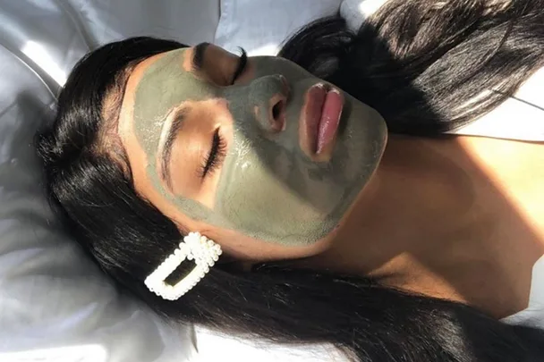 A woman with dark hair and pearl hair clip wears a green face mask, lying down on a white pillow.