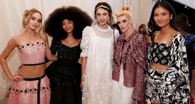 Group of five women in fashionable outfits, posing at an event with a cream backdrop.